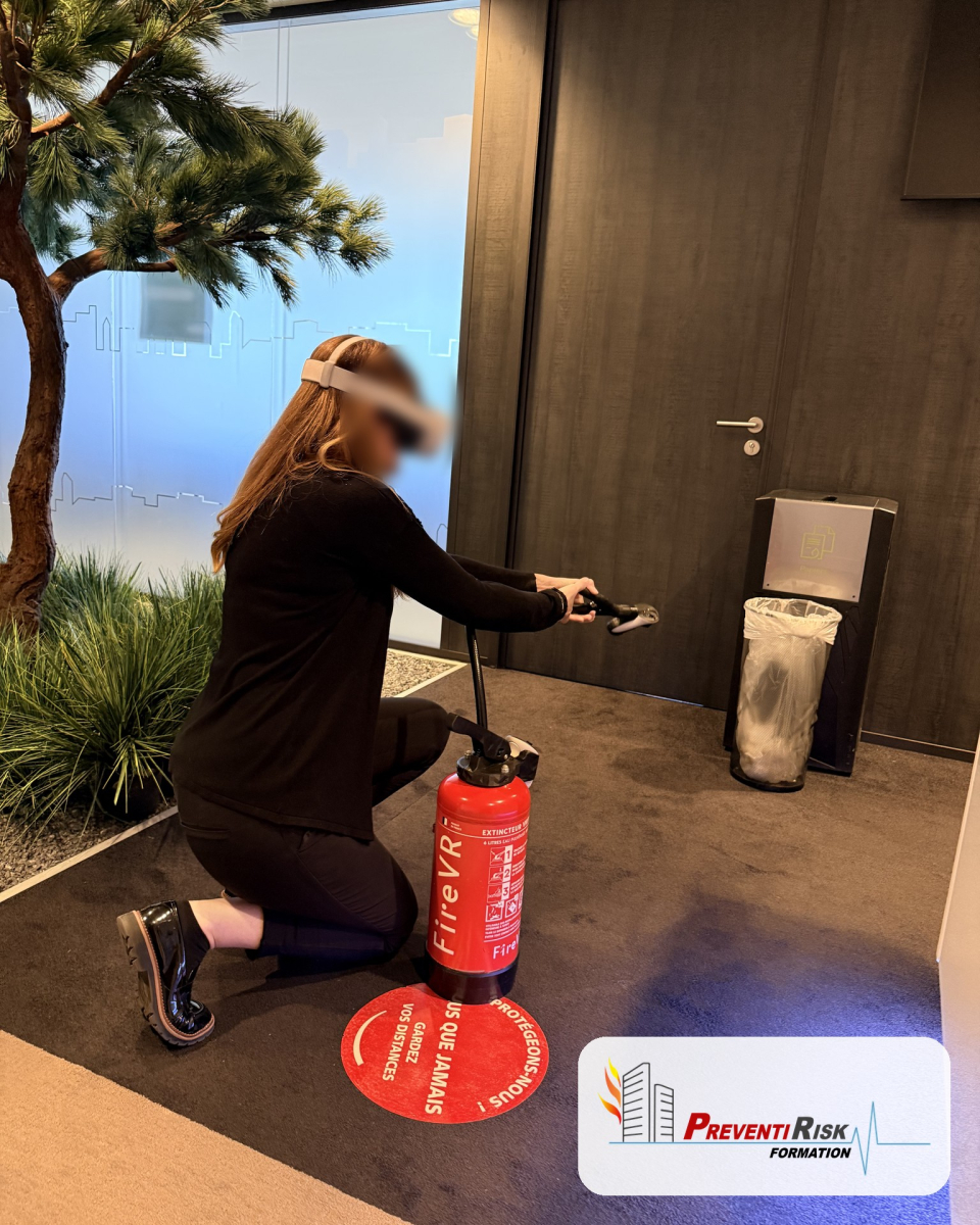 Une salariée, casque de réalité augmentée sur la tête, est à genou dans un couloir de bureaux. Elle dirige la lance d'un extincteur à eau vers un feu virtuel, lors d'une formation incendie PREVENTIRISK.